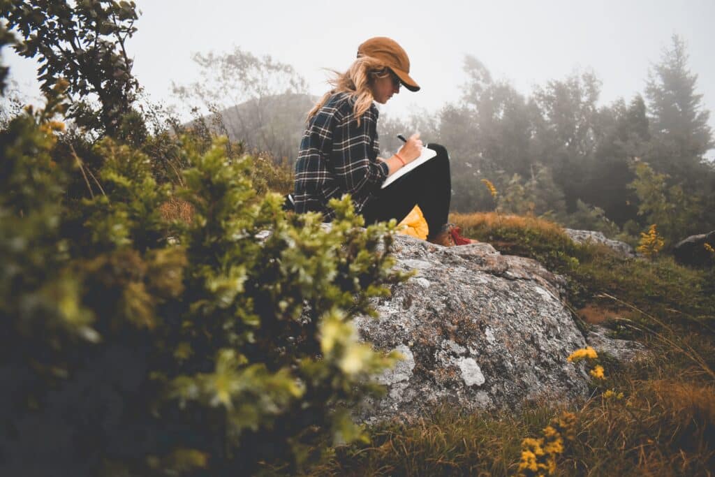 A woman sitting on a rock with grass and plants around her on a foggy day; she's writing in a journal.