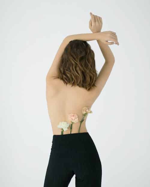 a woman without a top on facing away from the camera with flowers tucked into her waistband