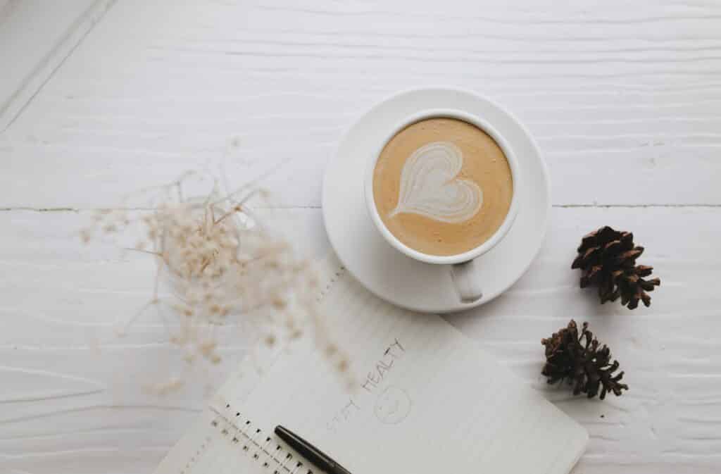 A coffee mug with a heart design depicted in coffee foam; pinecones; and a notepad and pan on a table