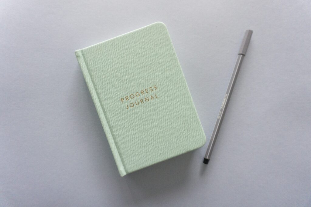 A closed journal and pen on a desk; the journal's cover says progress journal on it
