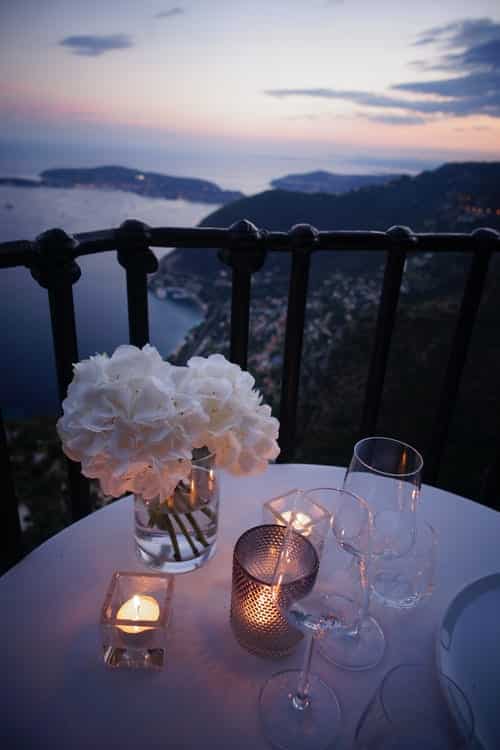 a plate, glasses, and flowers on a dinner table on a balcony overlooking a lake at dusk