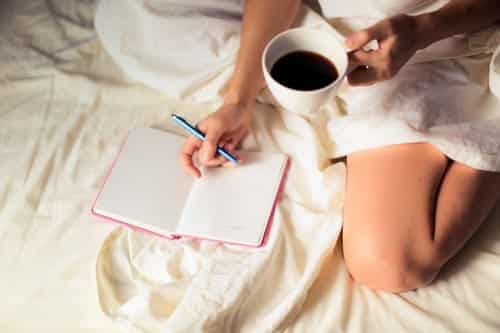 A woman wrapped in a duvet holding a mug of coffee and writing in her journal in bed