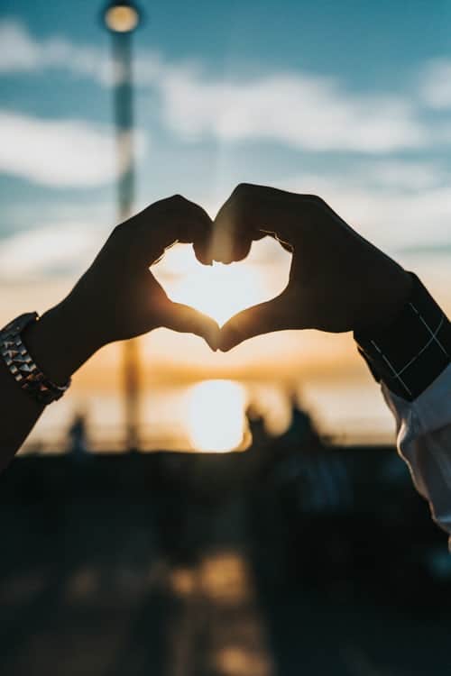 two hands making a love heart shape in front of the setting sun