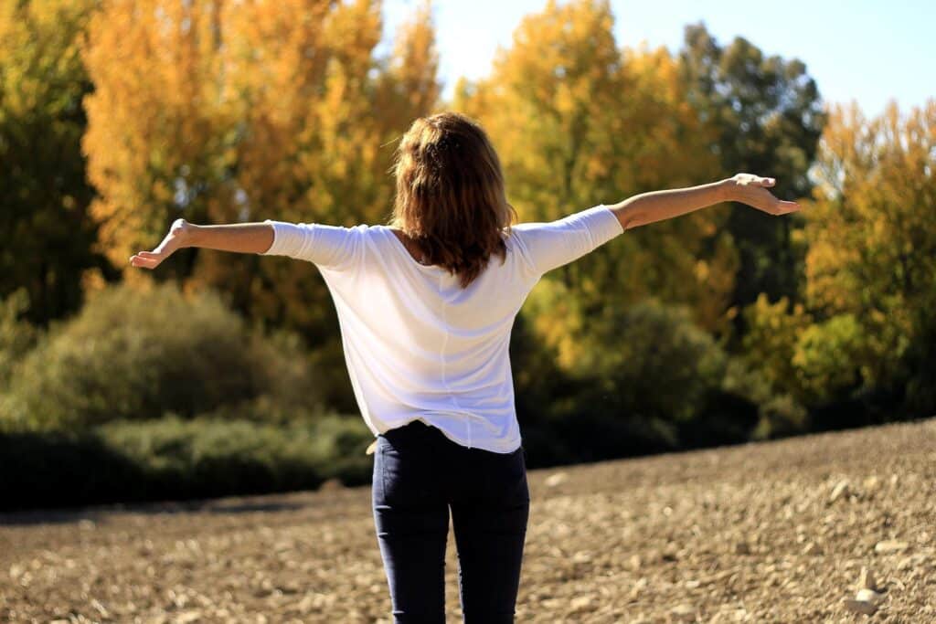 A woman with her arms outstretched standing in front of a forest with rays of sun beaming down on her.