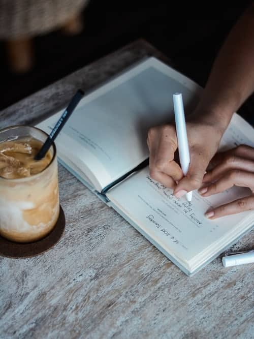 hands writing in a journal with an iced coffee next to them