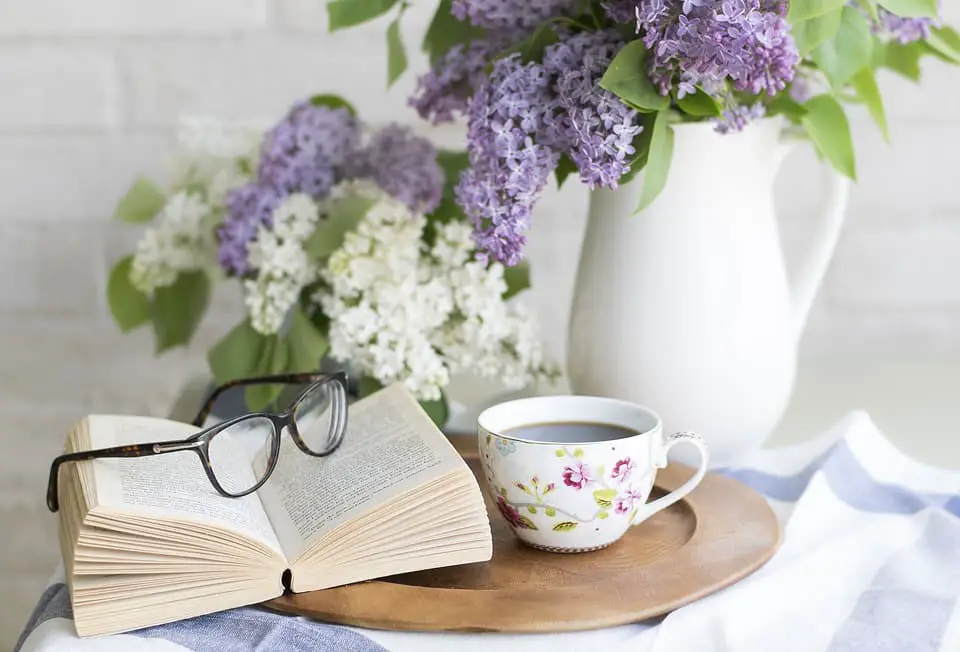 A journal, cup of tea, reading glasses, and vase with flowers on a table