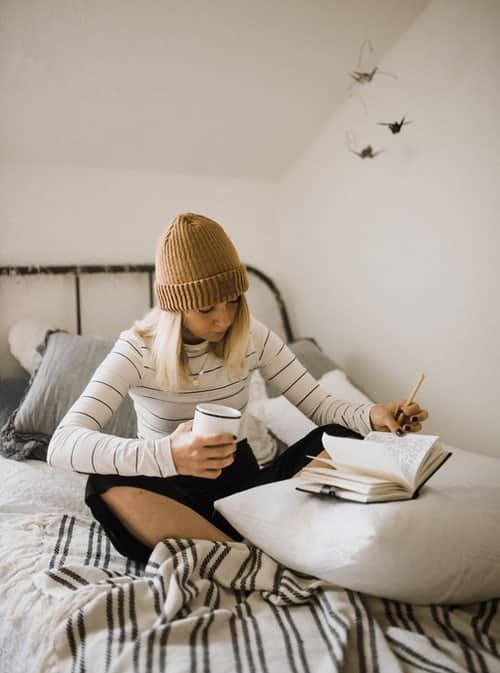 a woman sitting on her bed with a cup of coffee, writing in her journal