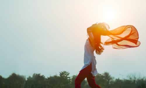 A woman standing in front of a sunny sky with her scarf blowing in the wind; her arms are raised skyward.