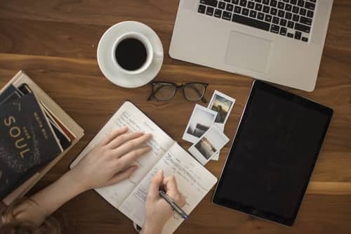 Person writing in a journal on a desk, with more journals, a coffee, and a laptop next to them.