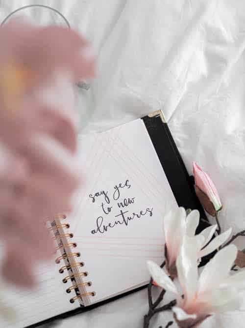 journal on a bed surrounded by flowers; the journal says "say yes to new adventures" on it