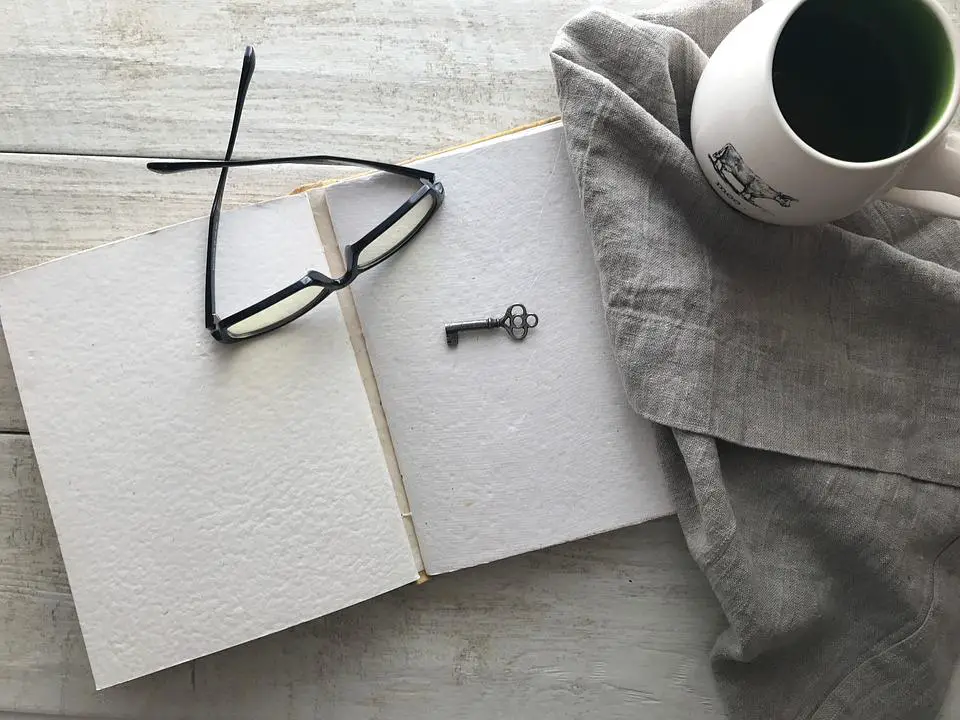An open journal with a key and glasses on top, next to a mug of coffee and a blanket