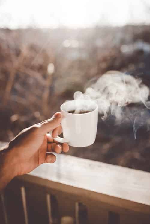 A hand holding a steaming cup of coffee over a porch