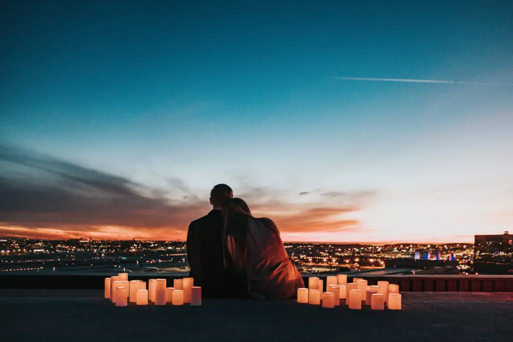 Two people learning against each other surrounded by candles; they're sat on the edge of a hill overlooking a town at sunset