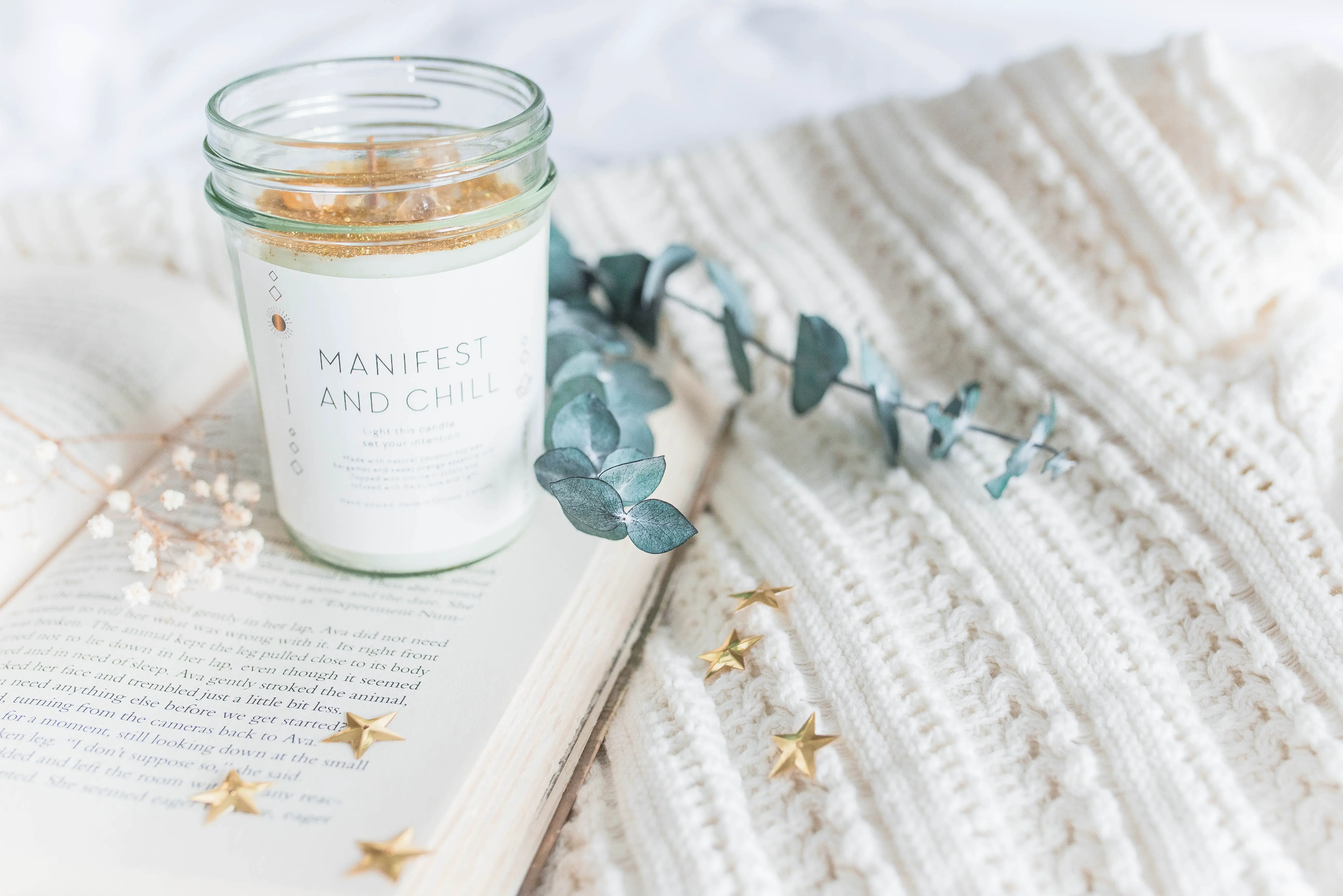 A white candle and leaves on a tablet cloth; the candle's jar reads, manifest and chill