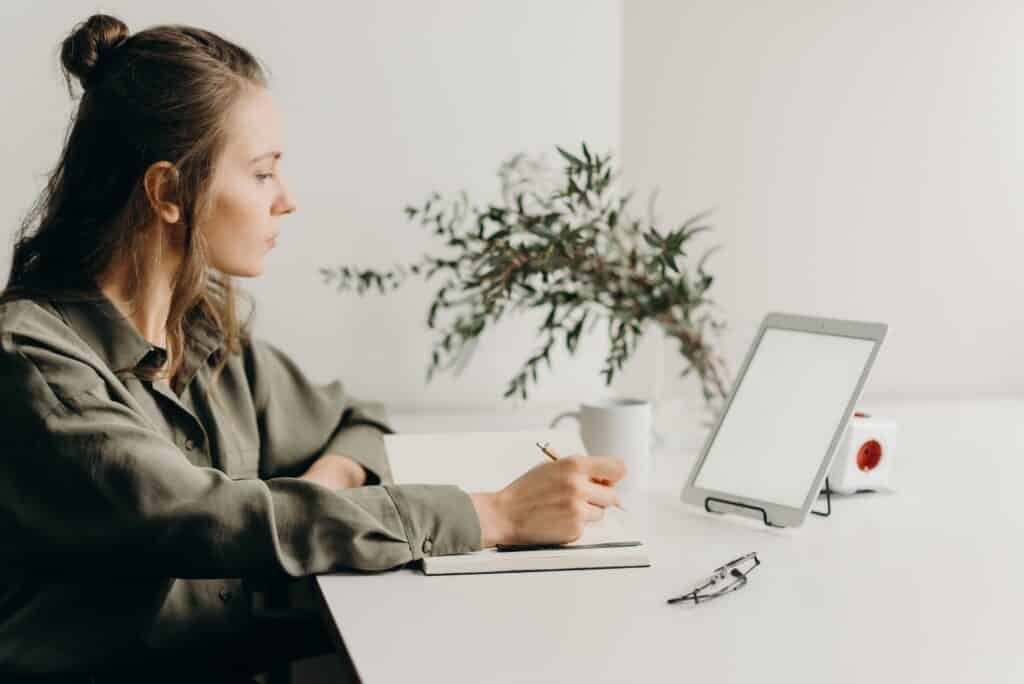 A woman sitting at her work desk with a writing pad, ipad, and mug of coffee.