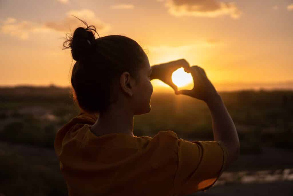 A girl making a heart-shape with her hands and holding it up to the sunset