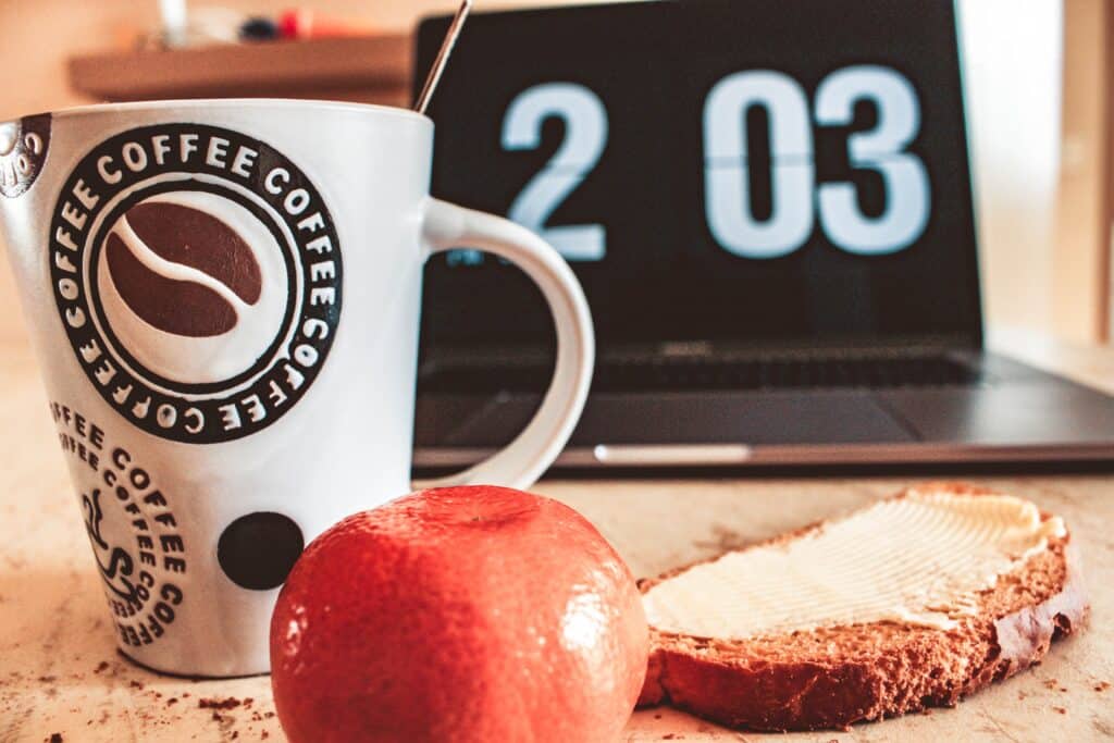A mug of coffee, slice of toast, and an apple in front of an open laptop.