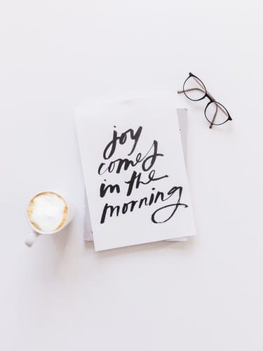 A piece of paper that reads "joy comes in the morning" on a desk alongside glasses and a cup of coffee