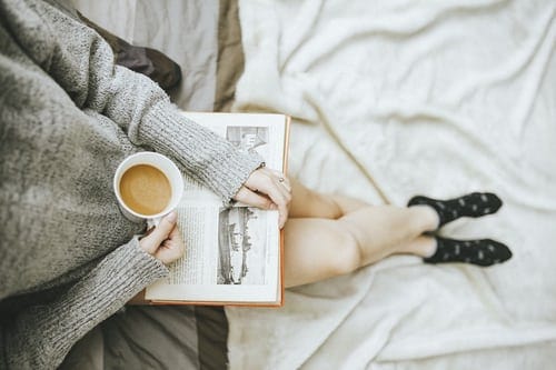 A person with crossed leg reading a book in her lap, holding a cup of coffee