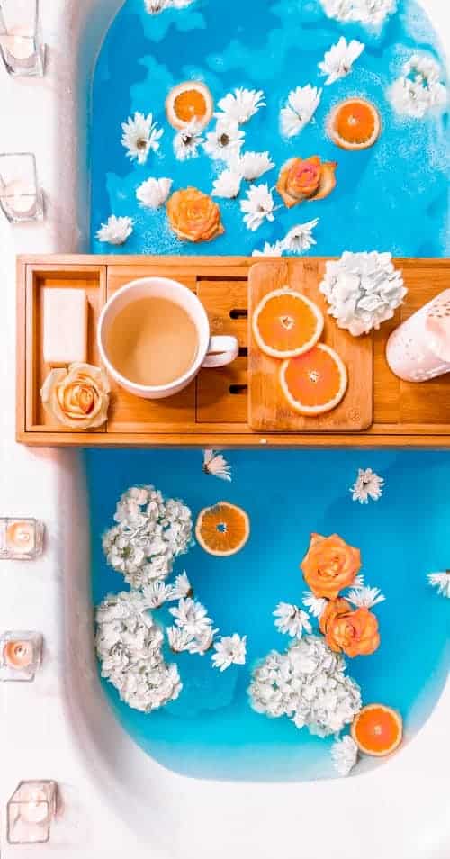 A cup of tea and sliced of orange on a bath tray; the bath water has flowers in it