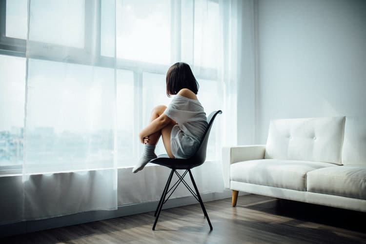 A girl sitting on a chair with her knees tucked into her chest staring out of a window