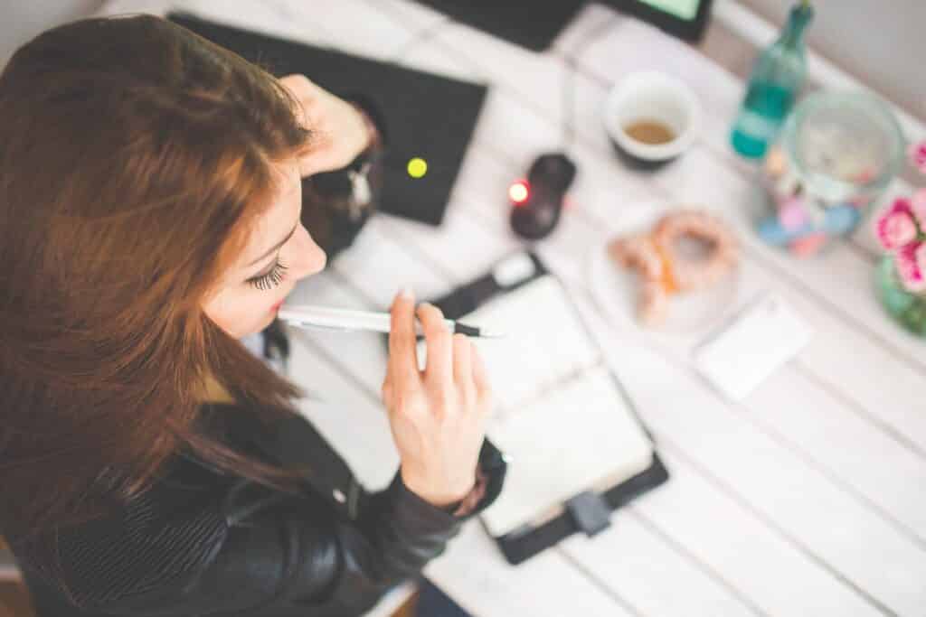 A young woman writing in planner at a desk; the desk has pastries, flowers and a laptop on it