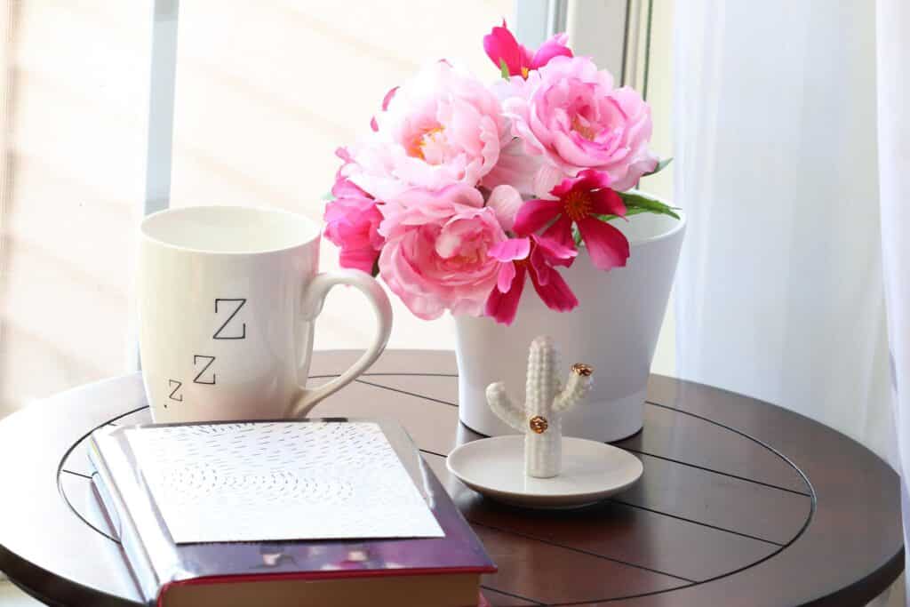 A journal, mug, and flowers on a small round table next to a window.